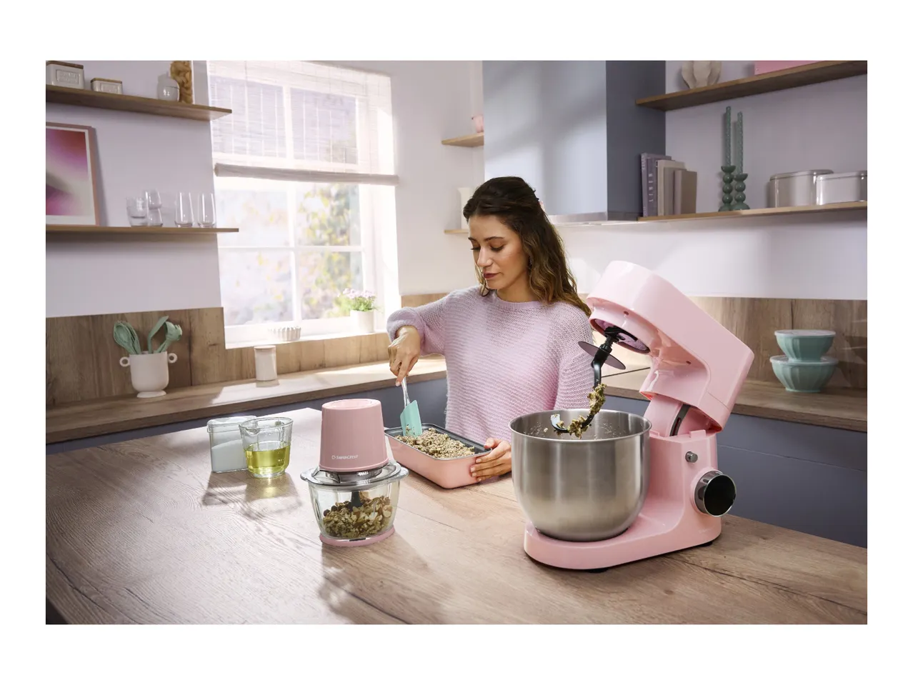Woman baking with a pink stand mixer and food chopper on a wooden kitchen counter.