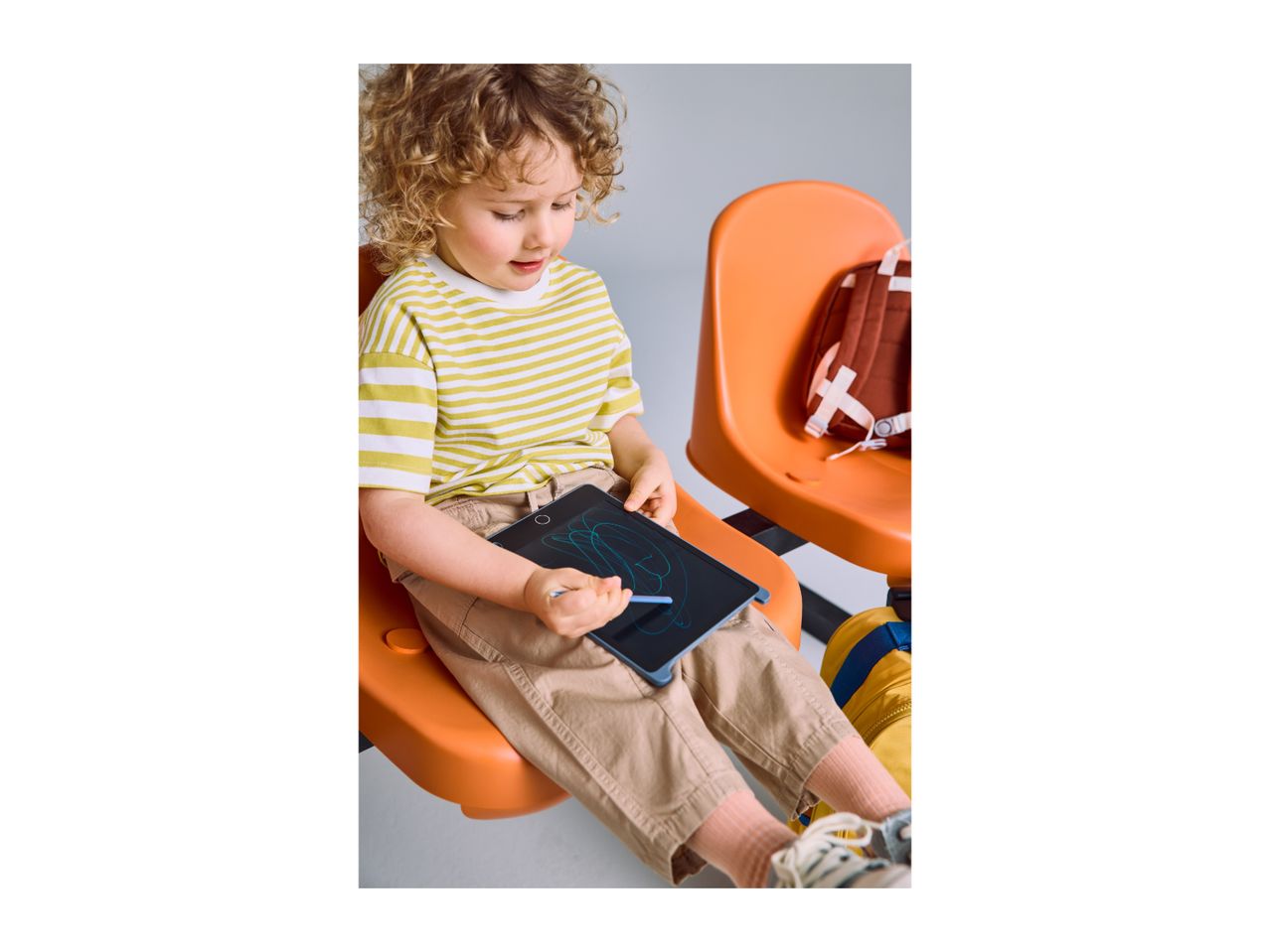 Child drawing on a digital writing tablet while sitting on an orange chair.