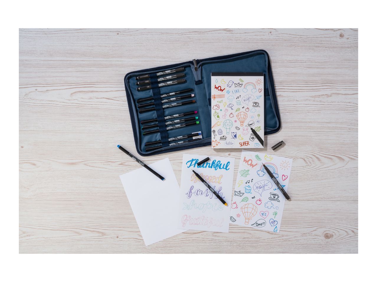 A set of colorful pens and markers, with two open notebooks featuring doodles and calligraphy.