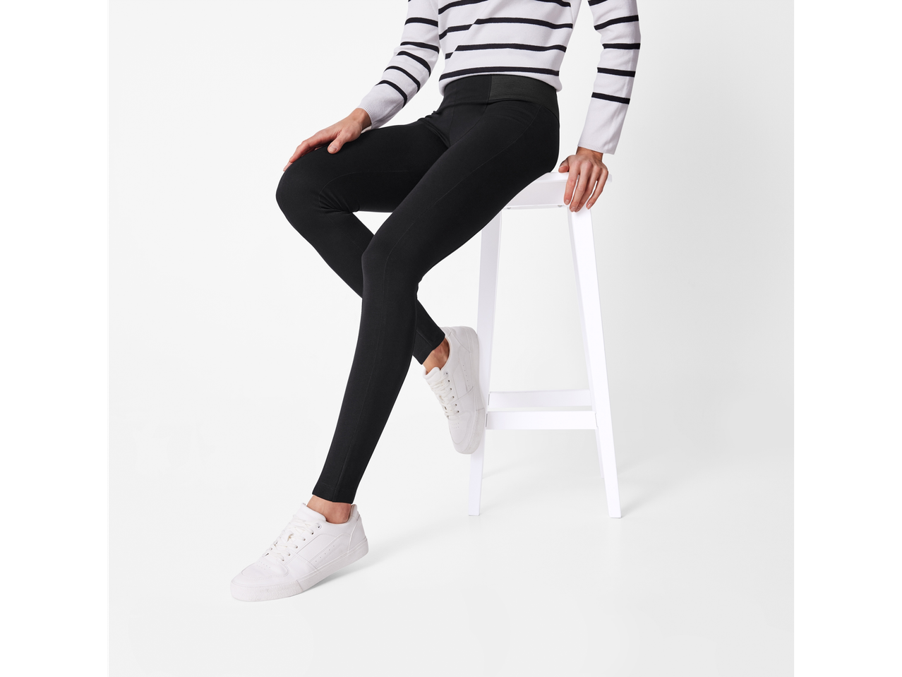 A woman in a striped sweater and black leggings sits on a white stool.