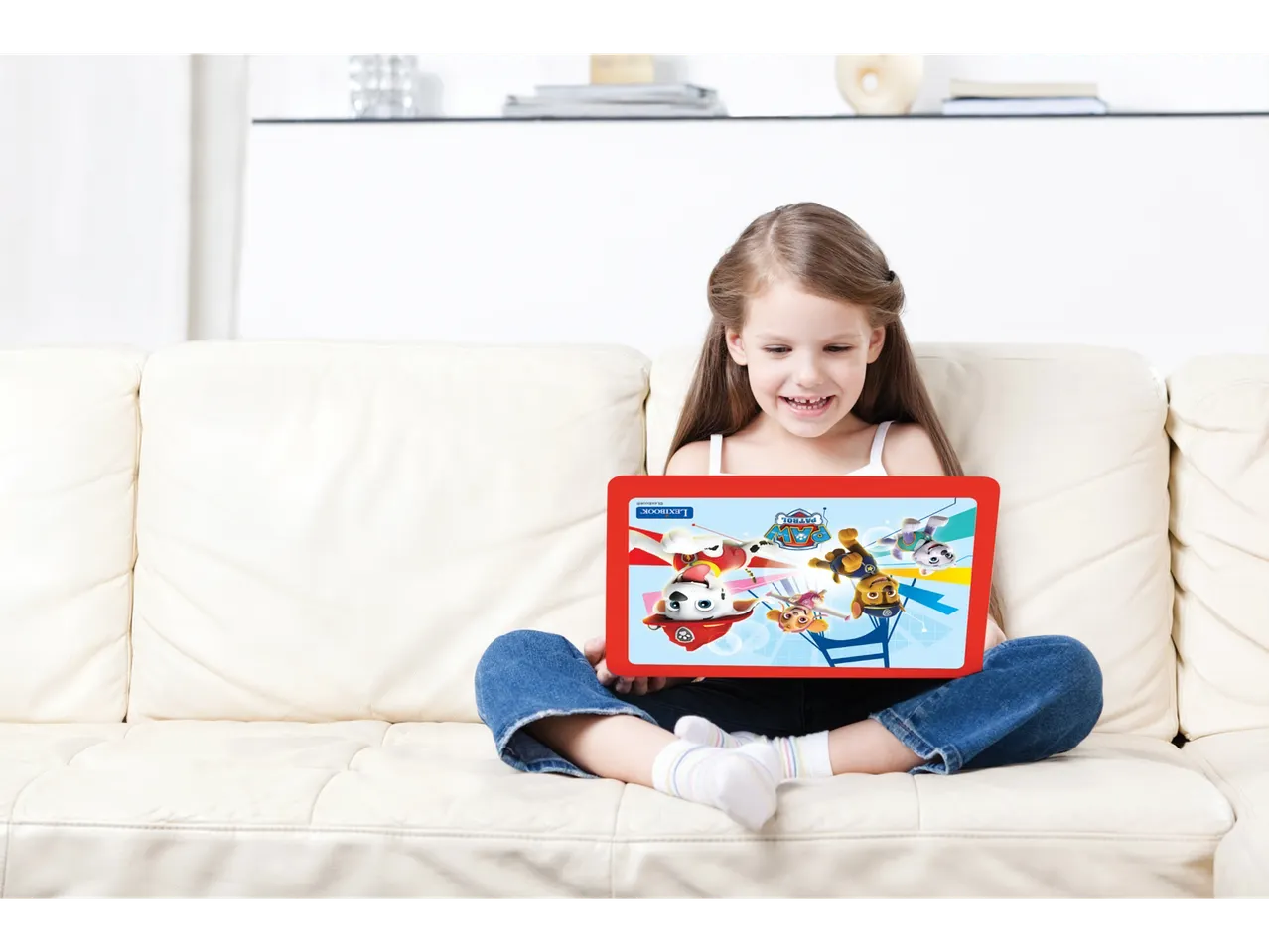 O fetiță zâmbitoare se uită la un laptop roșu cu tematică Paw Patrol pe canapea.