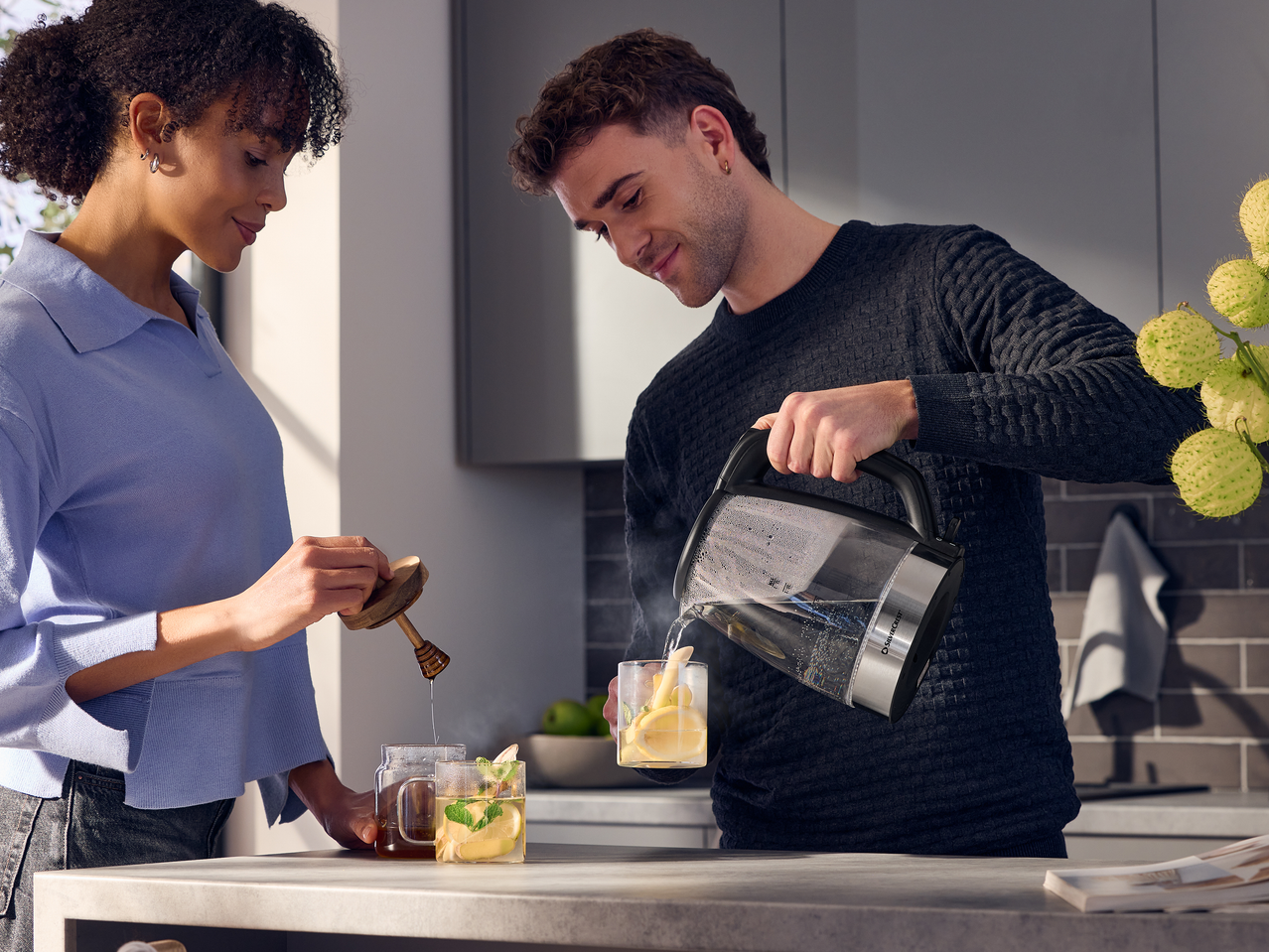 Ένα ζευγάρι ετοιμάζει ζεστά ροφήματα με έναν γυάλινο βραστήρα, μέλι και φρέσκα υλικά.