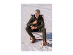 A smiling man in a dark green tracksuit and brown boots sits on a deck chair in the snow.