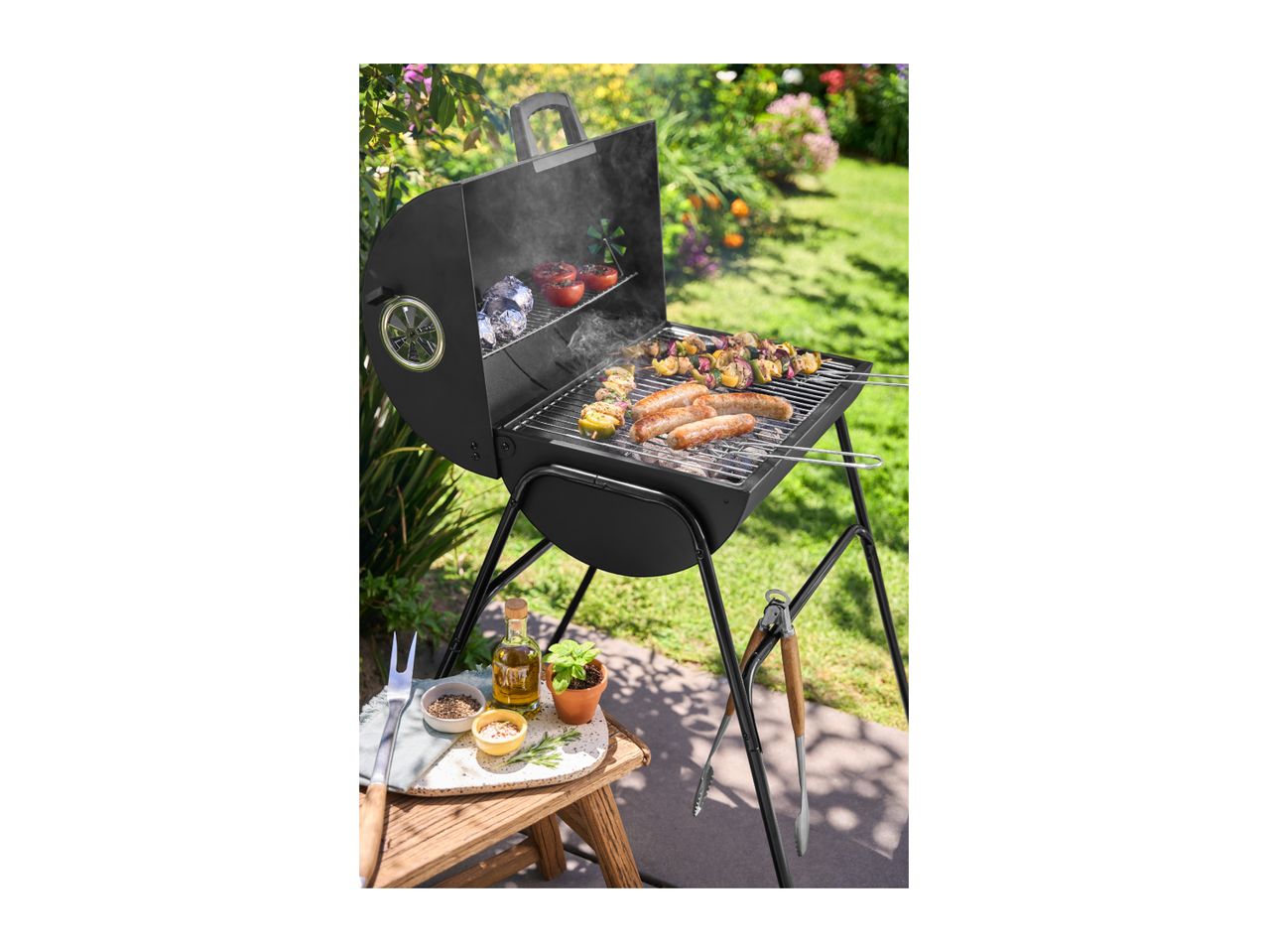 Black charcoal grill with sausages, vegetable skewers, and grilled vegetables in a garden setting.