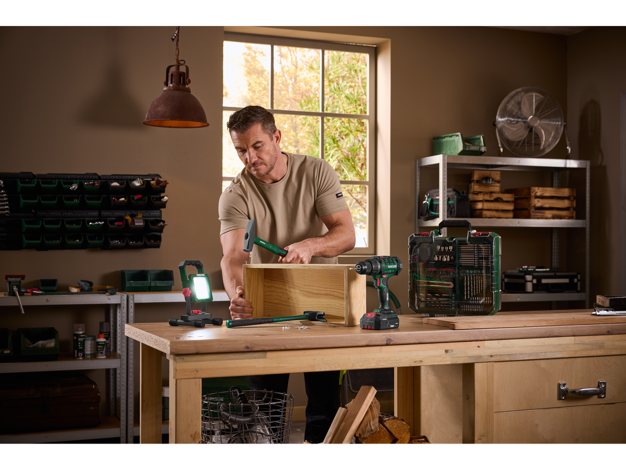 Un bărbat asamblează o cutie din lemn cu un ciocan, având alături o bormașină și un set de accesorii.