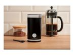 Black coffee grinder, French press, and ground coffee jar on a wooden counter.