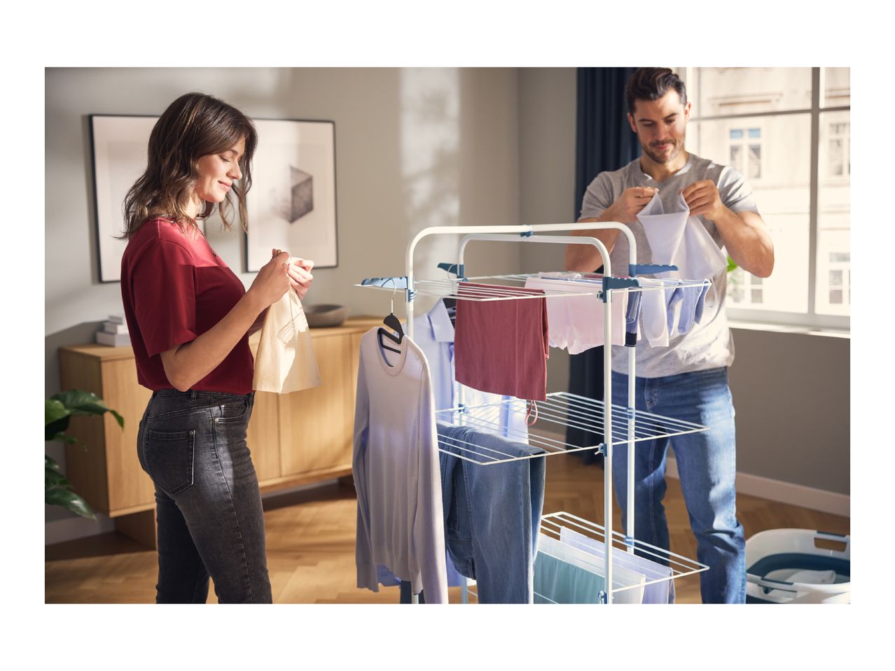 A couple hanging laundry on a white drying rack in a sunlit room.