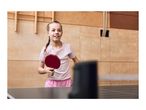 A young girl playing table tennis, holding a red paddle and smiling.