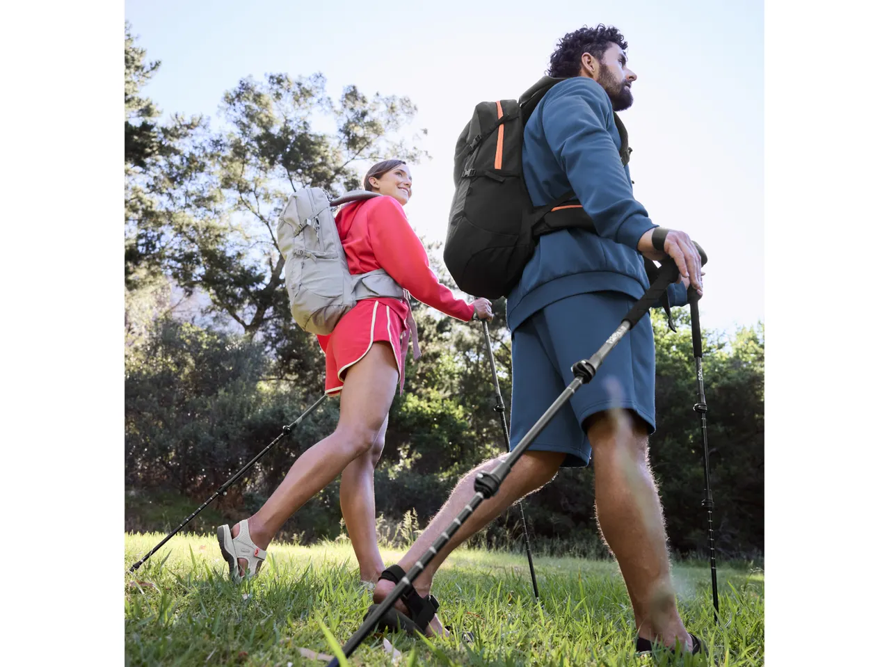 Two hikers with backpacks and trekking poles walk through a grassy, wooded area.