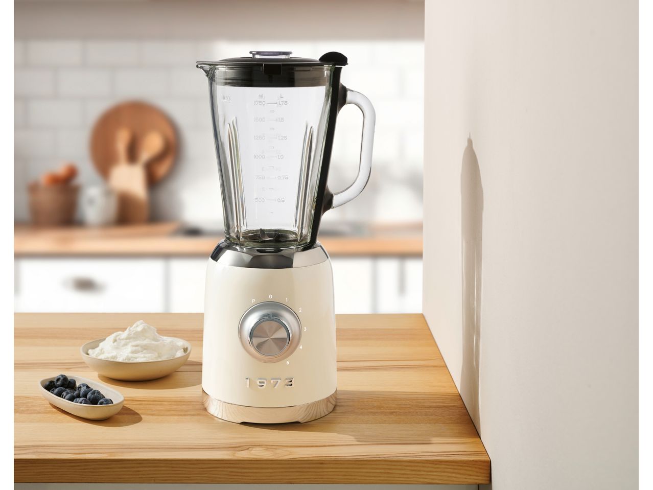 A retro-style cream blender with a glass jar on a wooden counter, next to bowls of blueberries and cream.