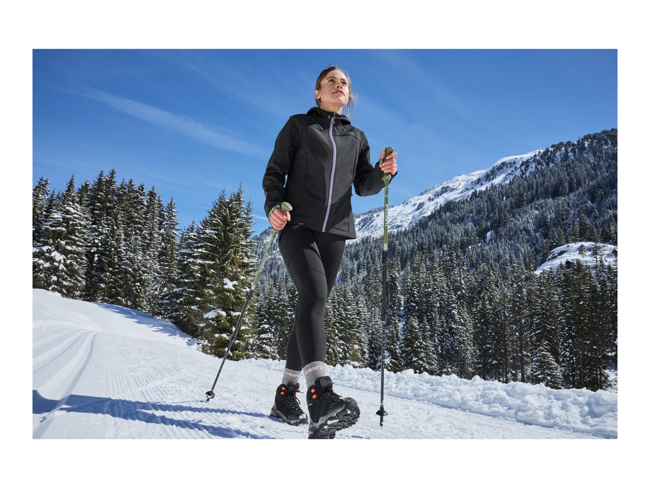 A woman winter hiking on a snowy trail with trekking poles, wearing a black jacket and leggings.