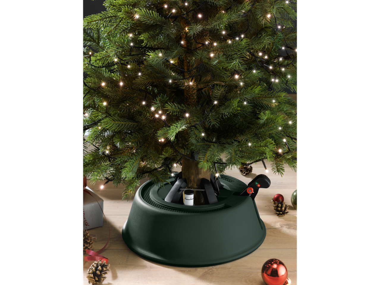 LIVARNO home Christmas Tree Stand in dark green, holding a lit Christmas tree.