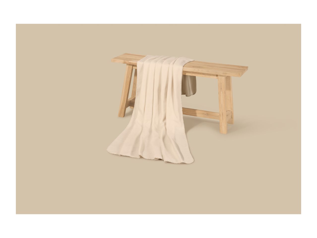 A cream-colored blanket draped over a light wooden bench on a neutral background.