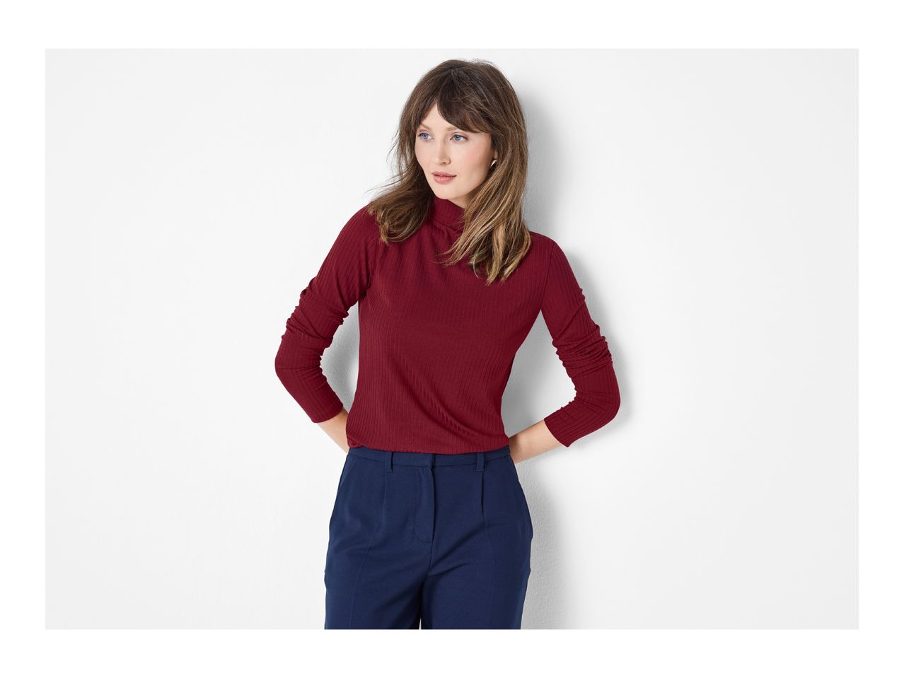 Woman in a red ribbed turtleneck and navy blue trousers, hands on hips, looking left.
