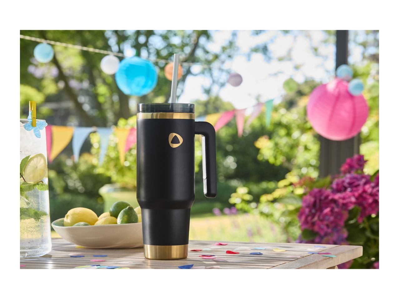 Black and gold travel mug with a straw on a wooden table with drinks and party decorations.