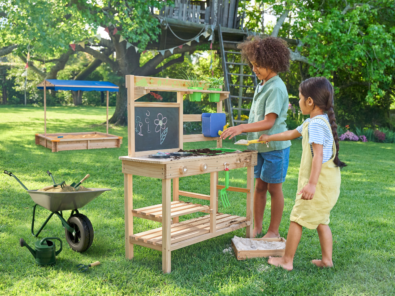 Две деца играят с дървена градинска маса за игра, пълна с пръст и инструменти, на зелена морава.