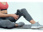 A person in athletic wear uses a black massage gun on their leg while sitting on a mat.