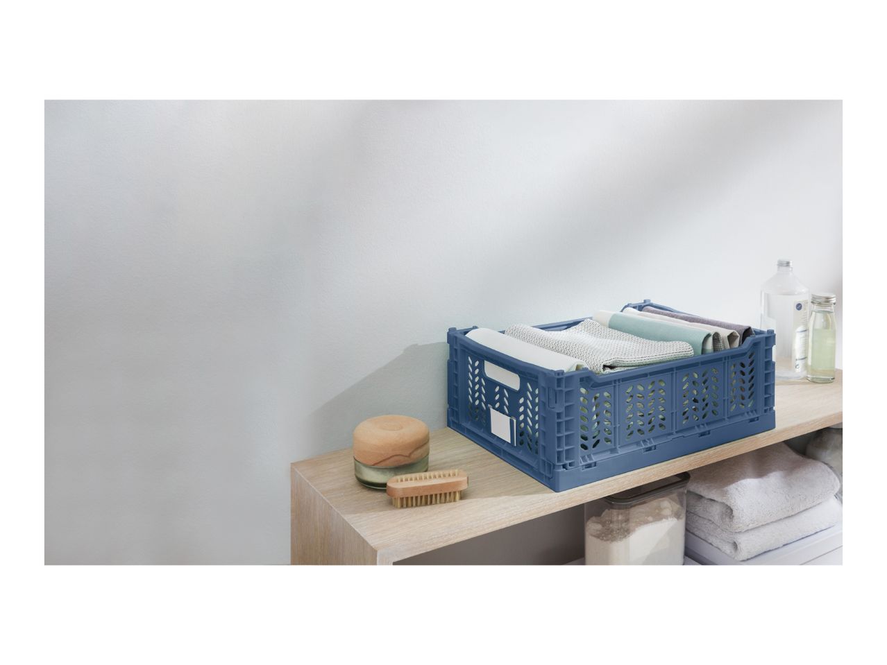 Blue foldable crate with textiles, a brush, and bottles on a wooden shelf.