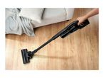 Person vacuuming a wooden floor with a cordless stick vacuum cleaner near a sofa.