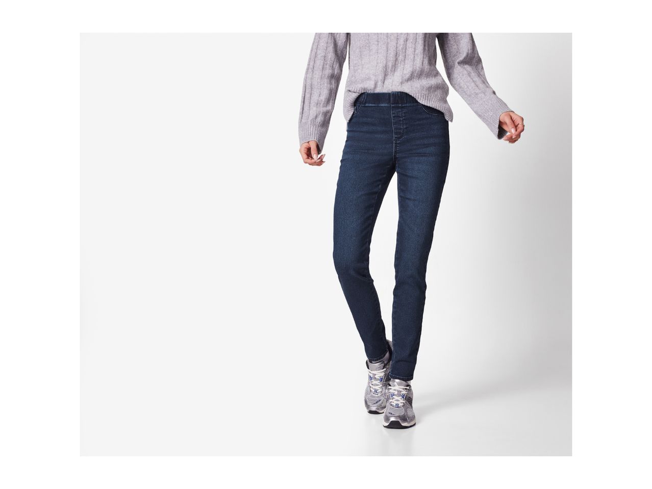 A person wearing a light gray ribbed sweater, dark blue jeans, and gray sneakers.