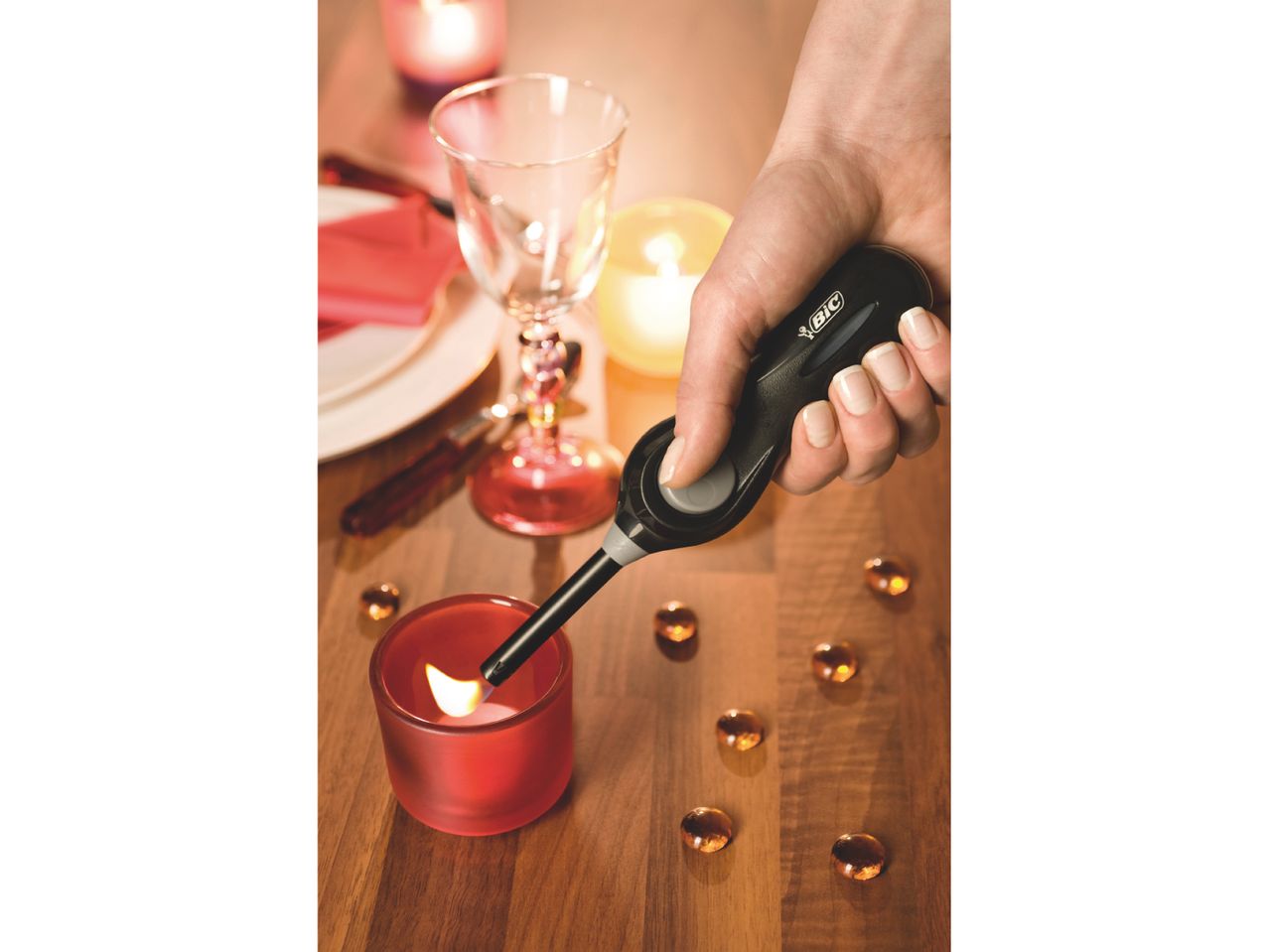 A hand holds a long lighter, igniting a candle among others on a wooden surface.
