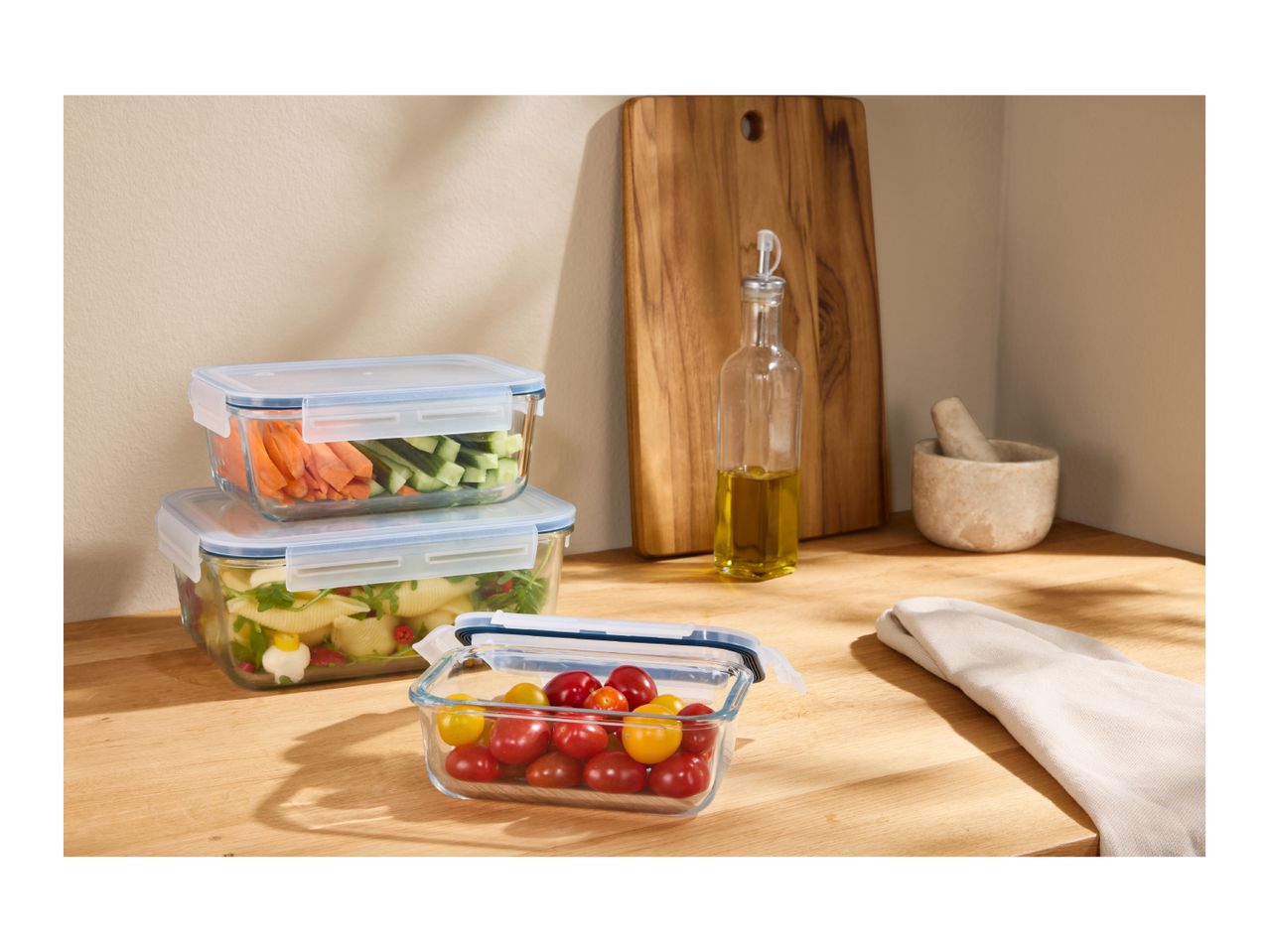 Glass food storage containers with fresh vegetables and pasta salad on a wooden kitchen counter.