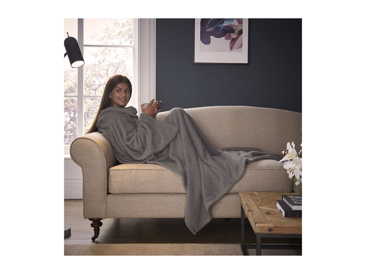 A woman in a grey hooded blanket on a beige sofa, holding a mug.