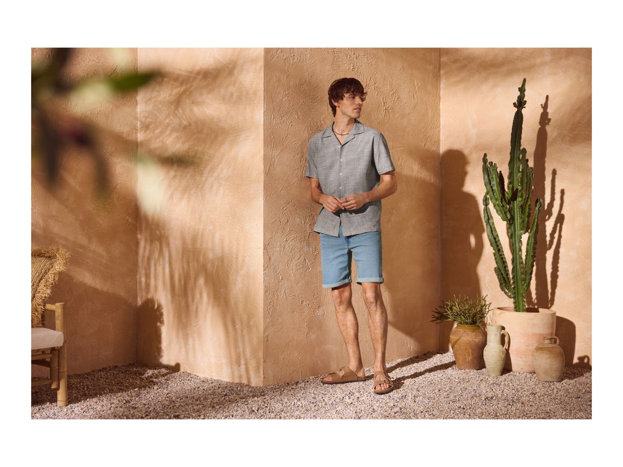 Man in a grey short-sleeved shirt, denim shorts, and sandals, standing in a sunny, desert-themed setting.