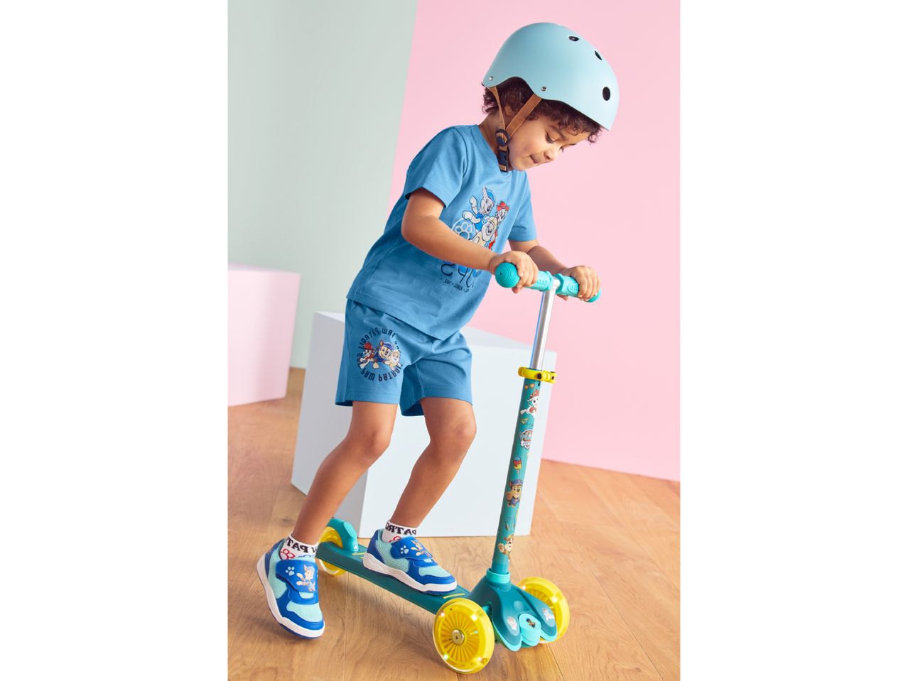 Boy in blue t-shirt and shorts, wearing a helmet, riding a scooter.