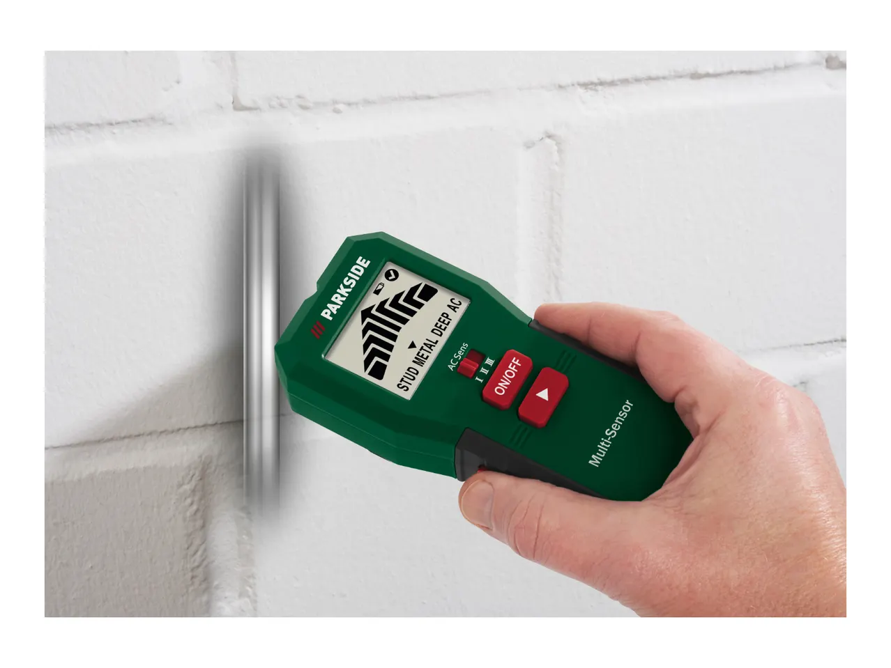 Hand holding a multi-sensor wall scanner detecting a pipe in a white brick wall.