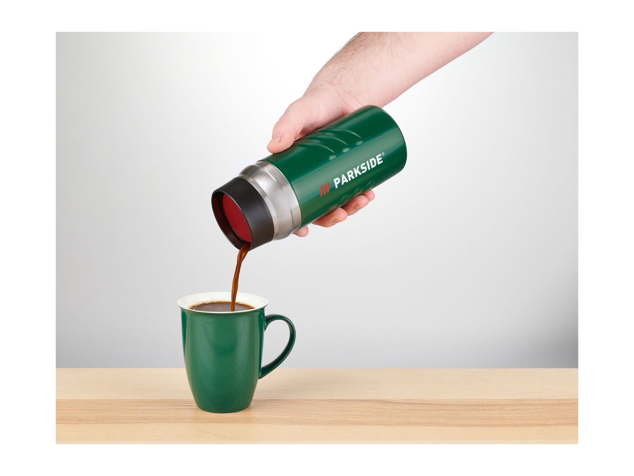 A hand pouring coffee from a green Parkside thermos into a green mug on a wooden table.