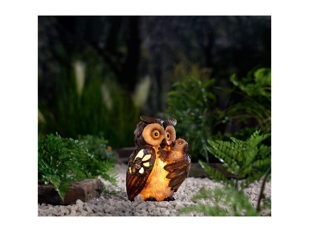 An illuminated owl garden ornament with a baby owl, surrounded by ferns and white pebbles at night.