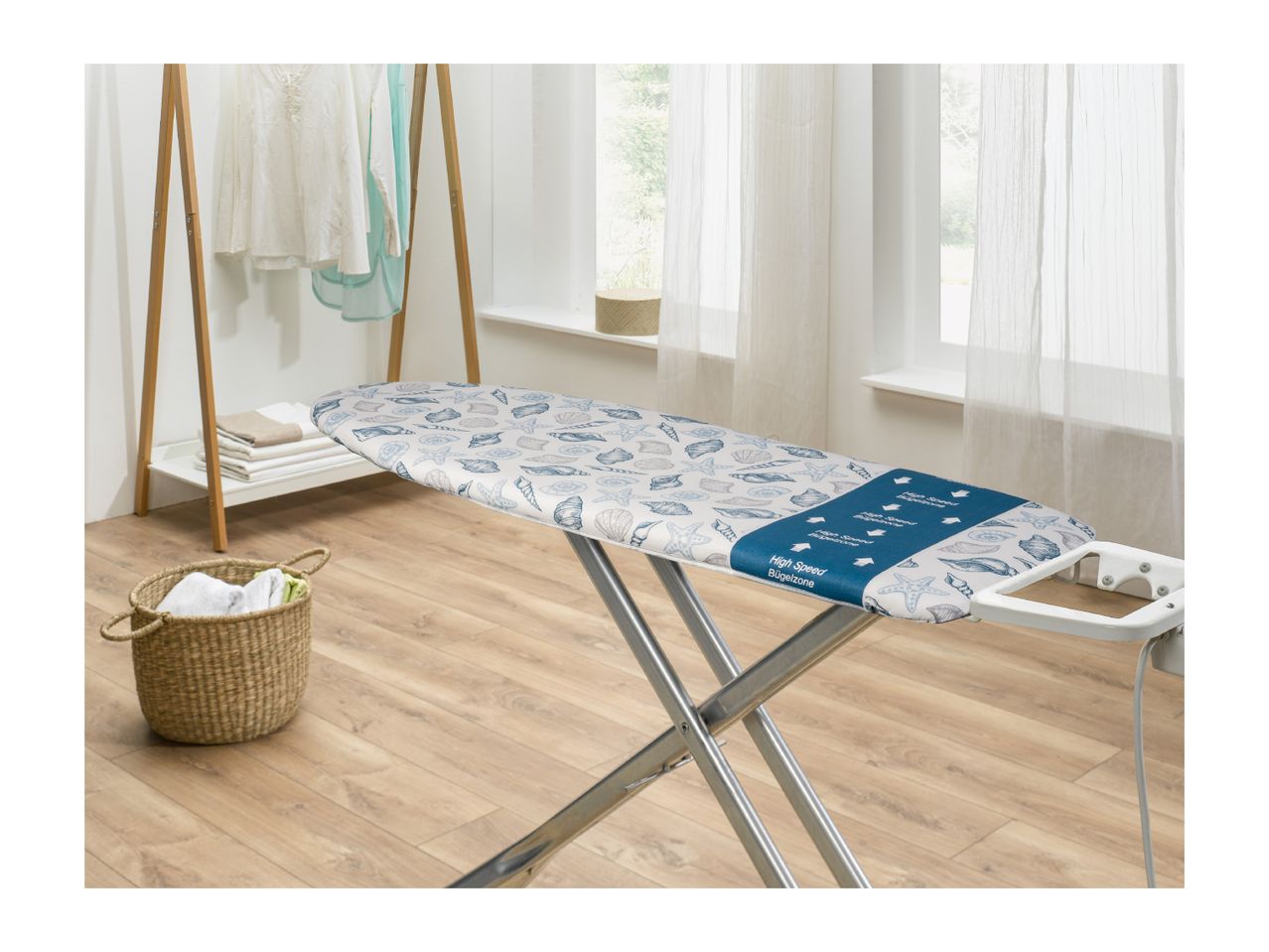Ironing board with seashell pattern, laundry basket, and clothes rack in a bright room.