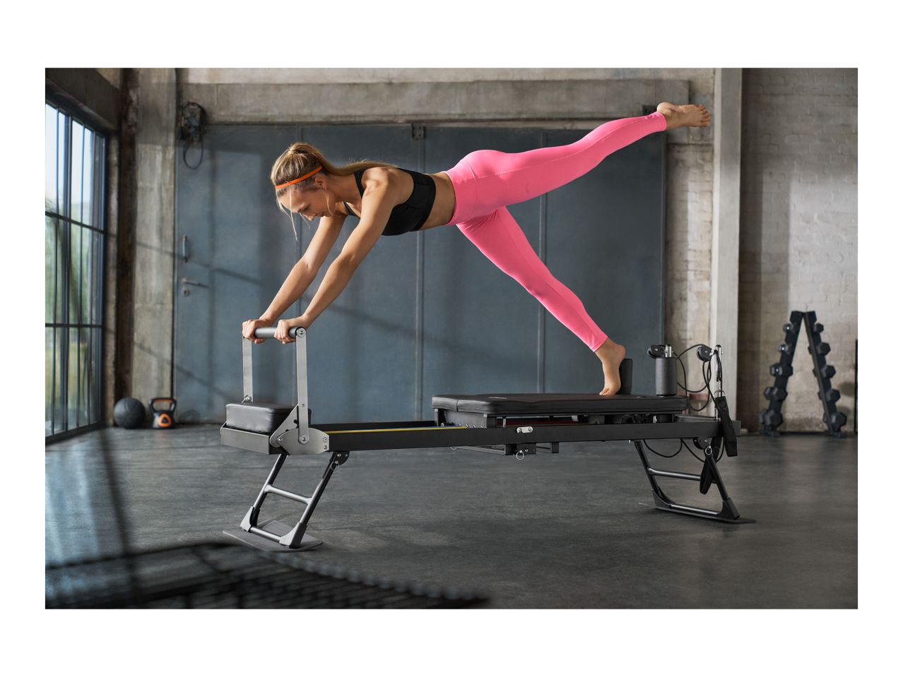 Woman in pink leggings and black sports bra doing pilates on a reformer machine.