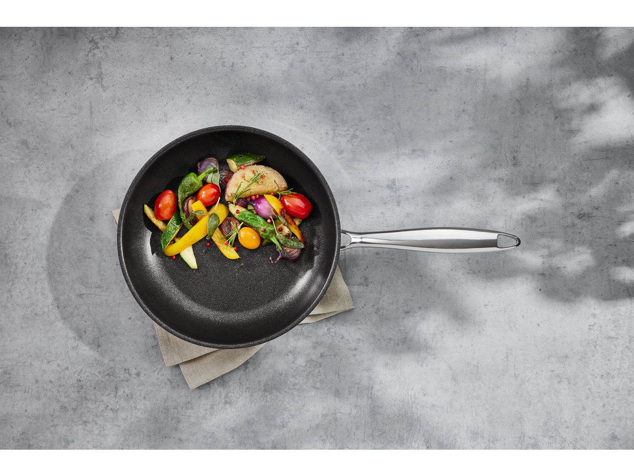 Tigaie cu legume proaspete, inclusiv roșii, ardei și ceapă, pe o suprafață gri.