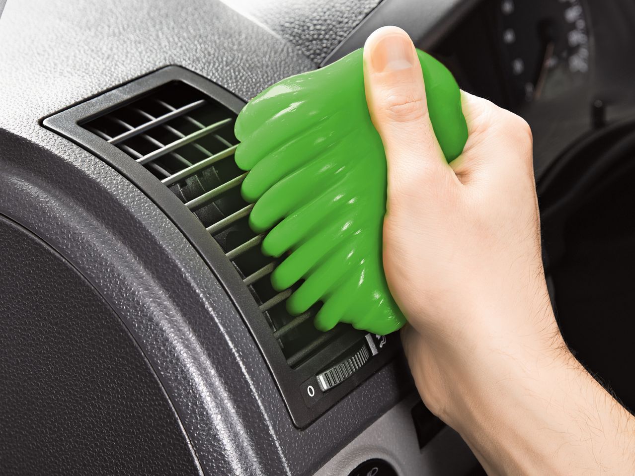 A hand uses green cleaning putty to clean a car's air vent.