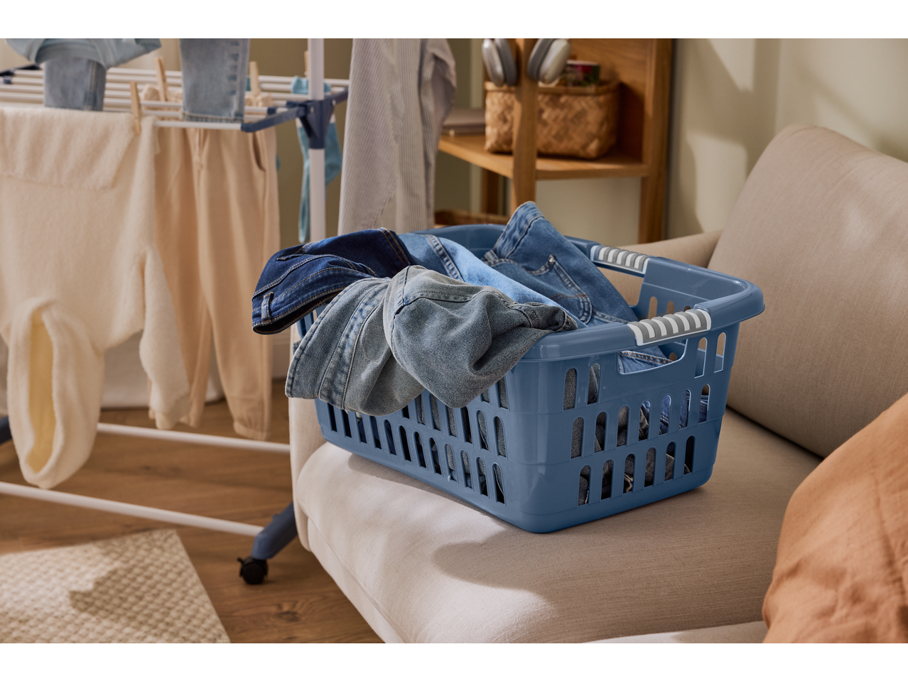 Blue laundry basket filled with denim clothes on a sofa, with a drying rack in the background.