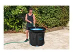 Man filling a black portable ice bath with water from a green hose in a garden.