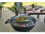 Vegetables cooking in a pan on a charcoal grill outdoors, with a set table in the background.