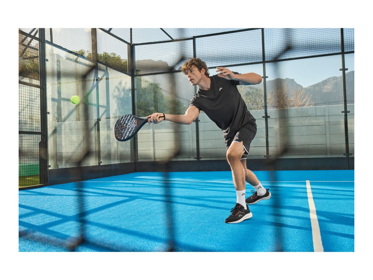 Man in black sportswear playing padel on a blue court, holding a racket, with a ball in the air.