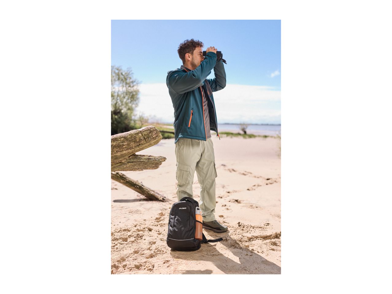 Man in outdoor jacket and cargo pants looking through binoculars on a sandy beach.