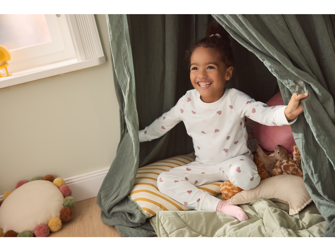 Happy child in Lupilu Kids' Pyjamas with pink hearts, sitting in a play tent.