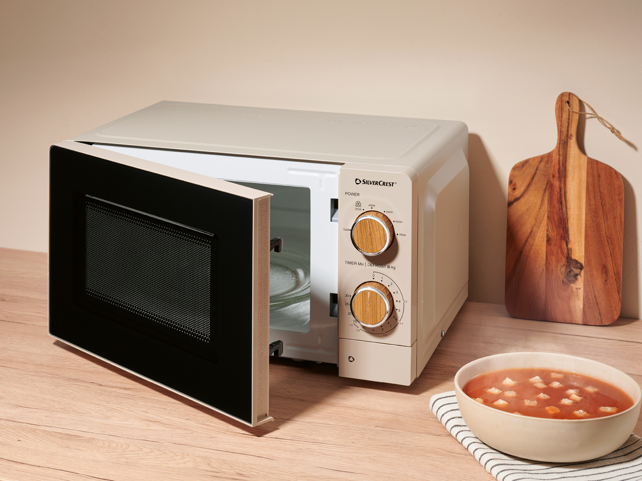 Бежова микровълнова печка с отворена врата, купа супа и дъска за рязане на дърво.