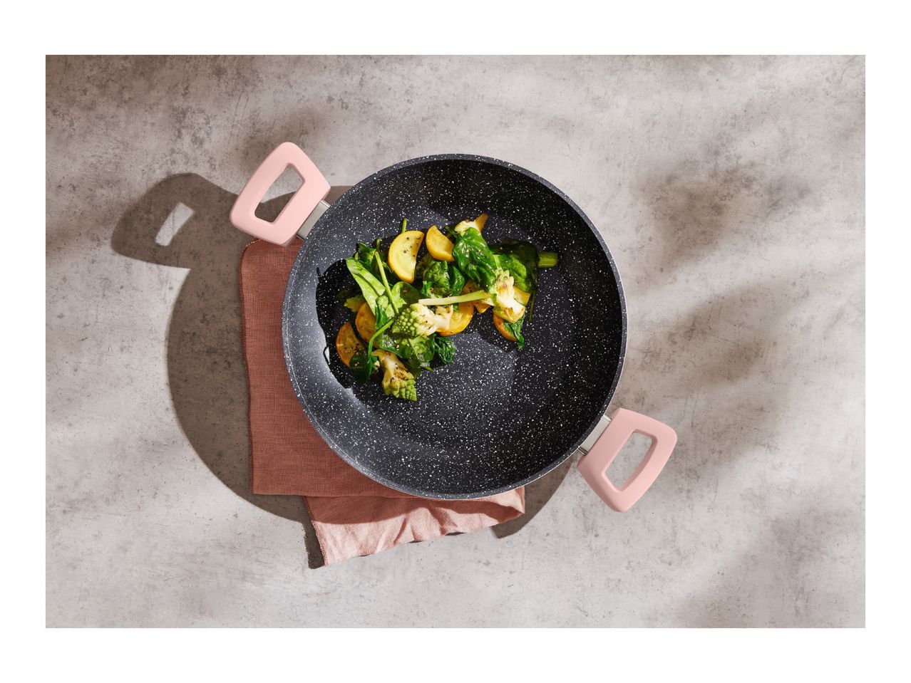 Overhead shot of a black speckled pan with pink handles, filled with cooked vegetables.
