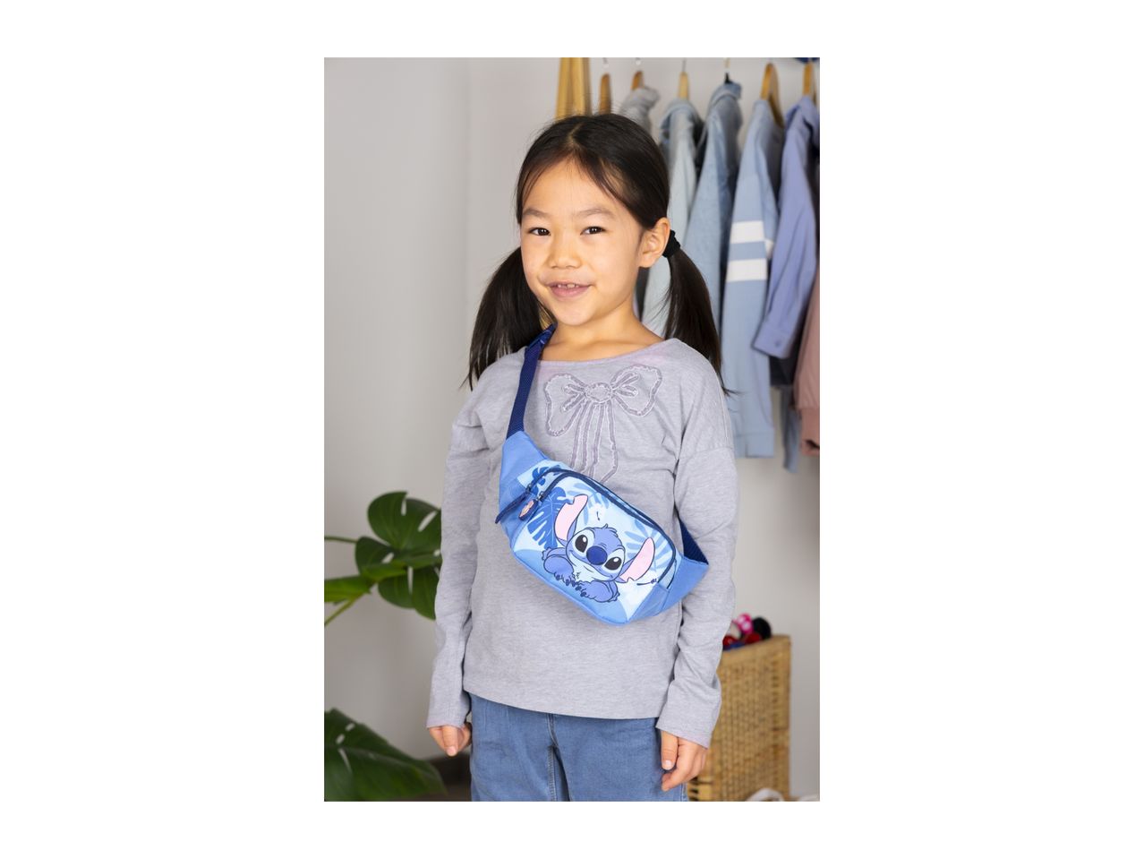 Smiling girl wearing a blue fanny pack with a cartoon character, standing in a room with clothes rack.