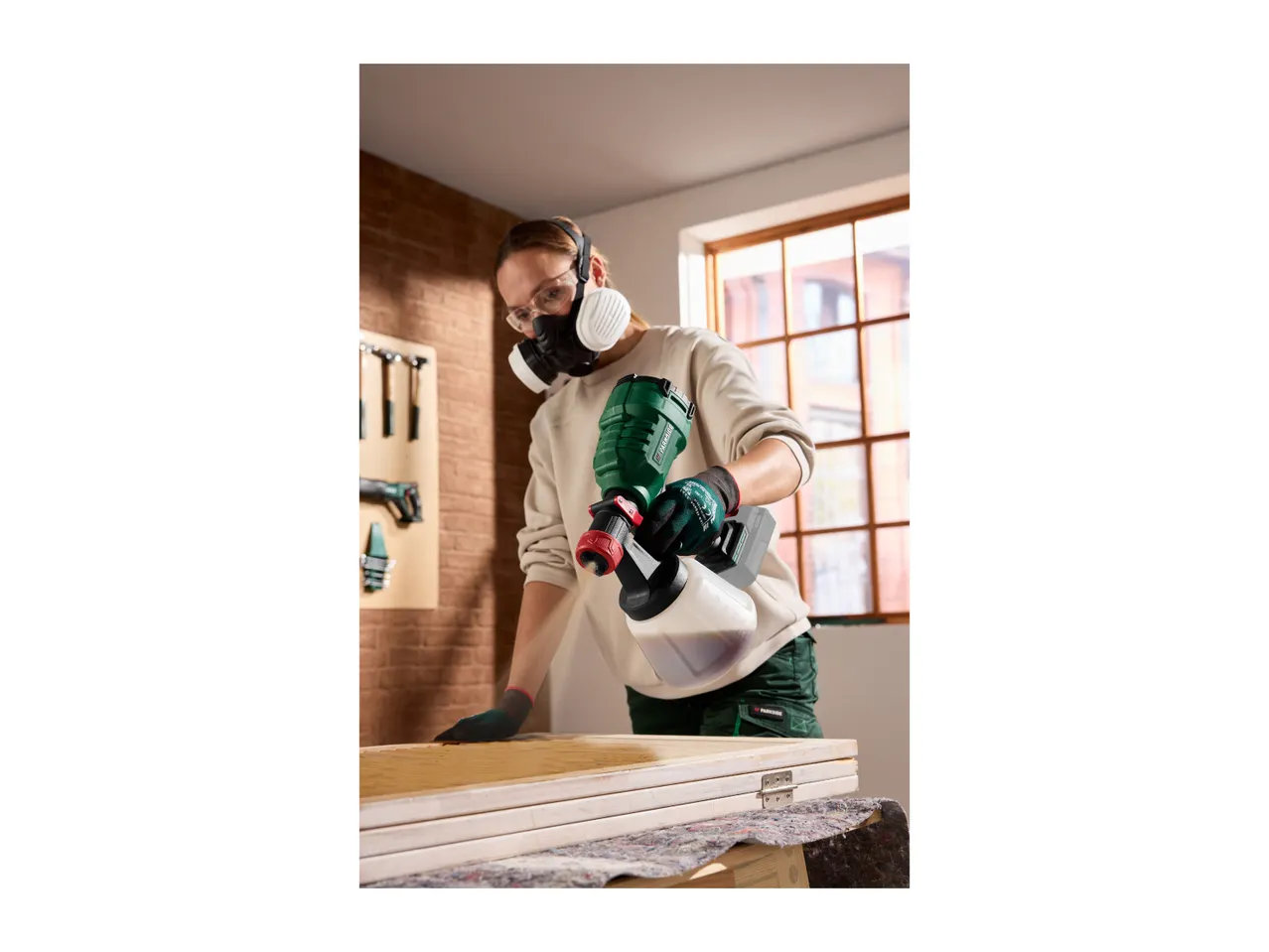 A person using a cordless paint sprayer to varnish a wooden surface.