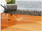 Cleaning a wooden deck with a pressure washer, with a planter and wall in the background.