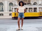 Woman in a cream top and denim shorts, standing on a street with a yellow tram passing by.