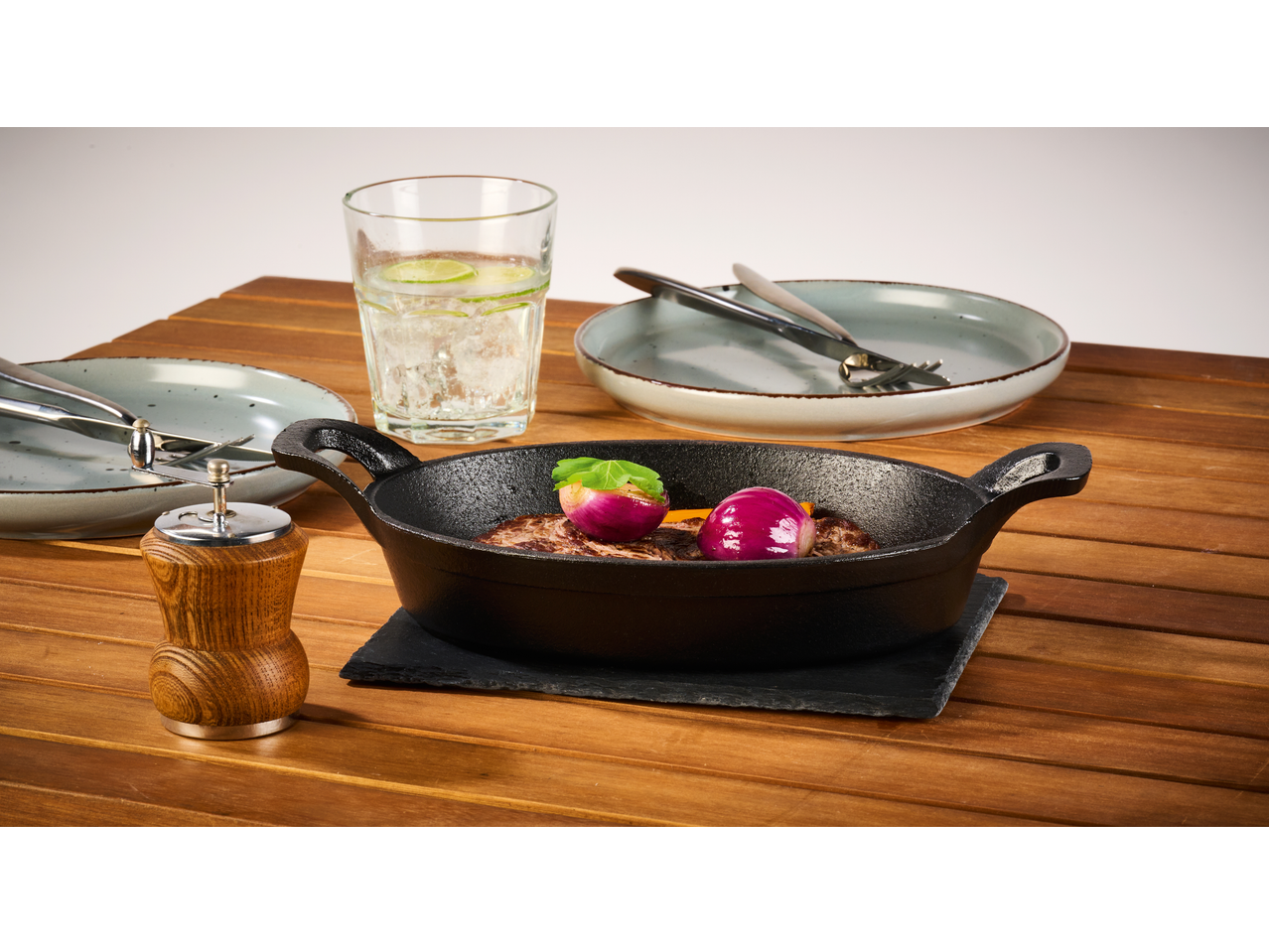 Tigaie din fontă cu carne și ceapă, pahar cu apă și farfurii pe o masă de lemn.