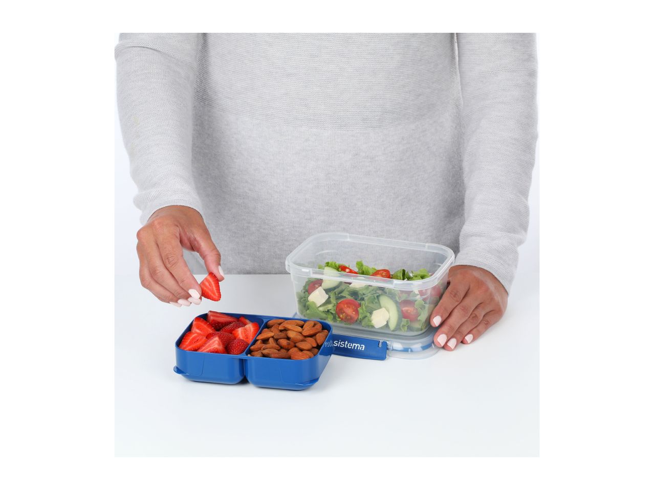 A person packing fresh strawberries, almonds, and salad into a blue and clear lunchbox.